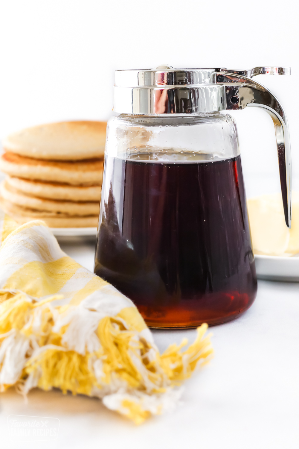 A jar of homemade maple syrup in front of a stack of pancakes and softened butter