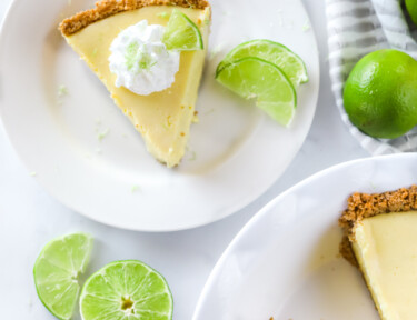 A slice of key lime pie with lime wedges, lime zest, and whipped cream topping