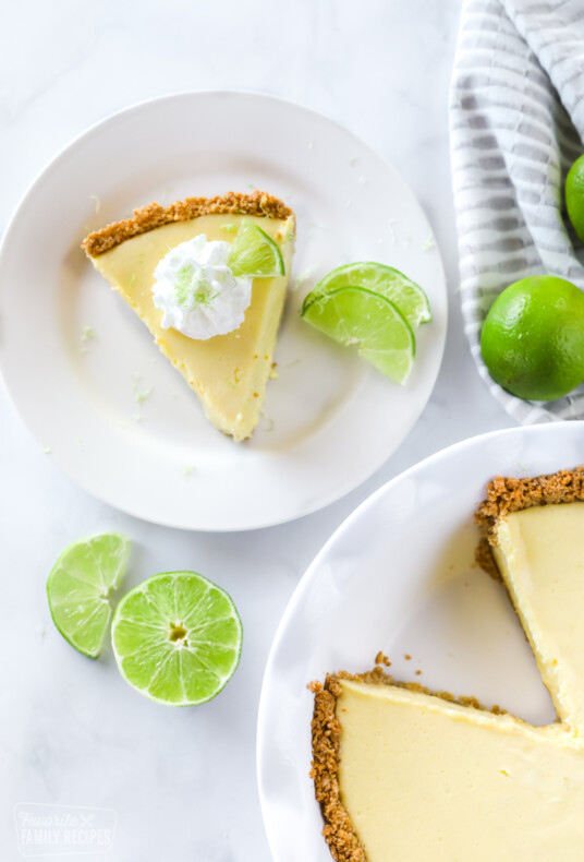 A slice of key lime pie with lime wedges, lime zest, and whipped cream topping