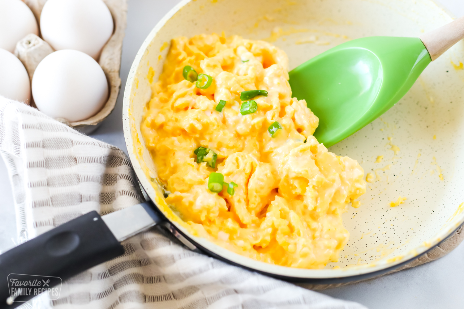 French style scrambled eggs in a pan with a green spatula. A carton of eggs is sitting next to the pan.
