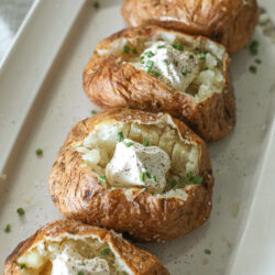 Four baked potatoes on a platter with sour cream and chives.