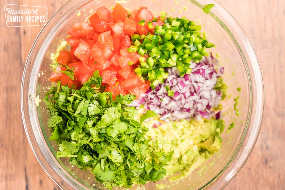 Mashed avocado, diced tomatoes, diced red onions, cilantro, and diced jalapeno in a large glass bowl