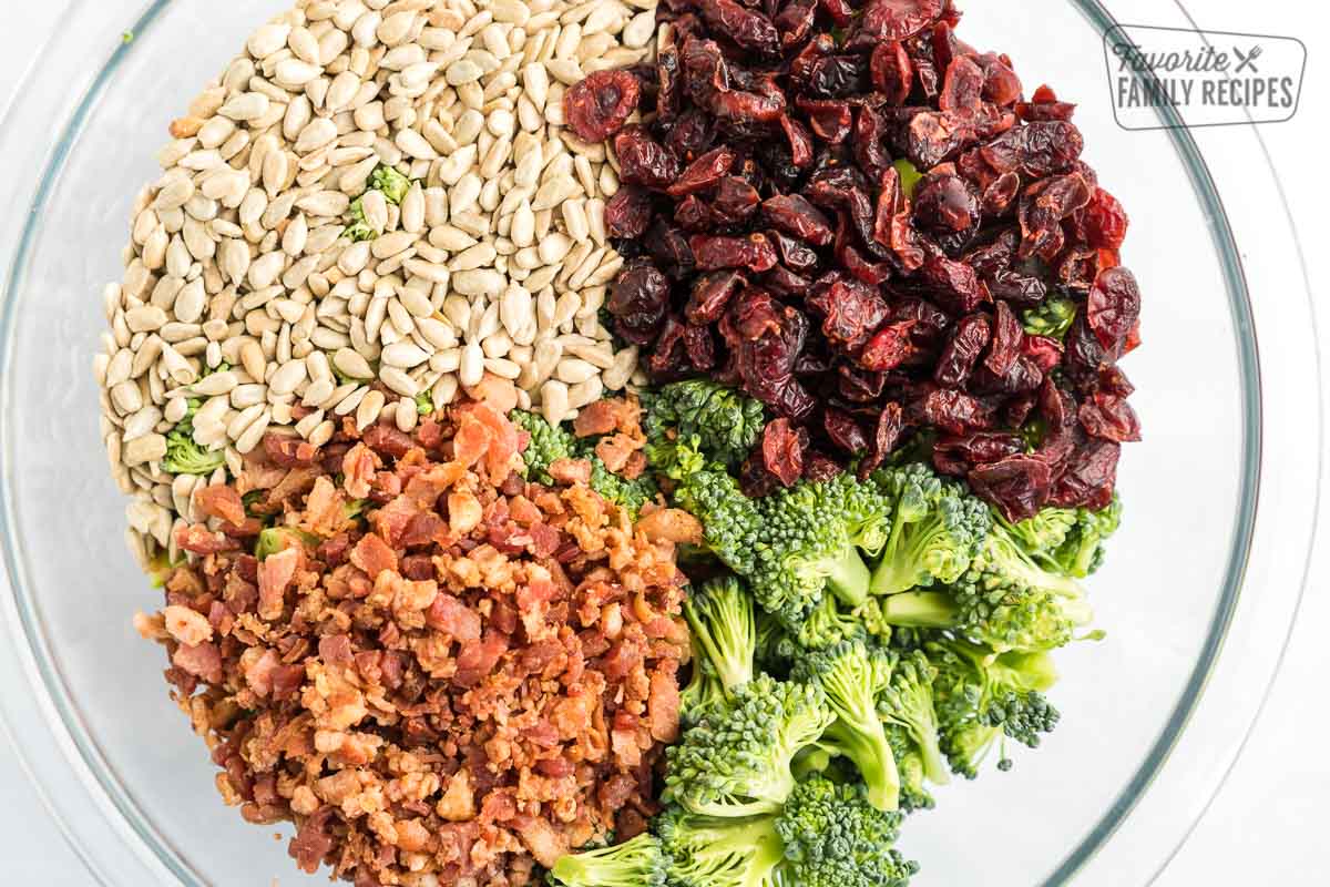 A large glass bowl full of broccoli, bacon bits, craisins, and sunflower kernels