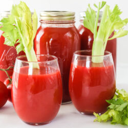 Two glasses of fresh tomato juice and canned tomato juice jars in the background.