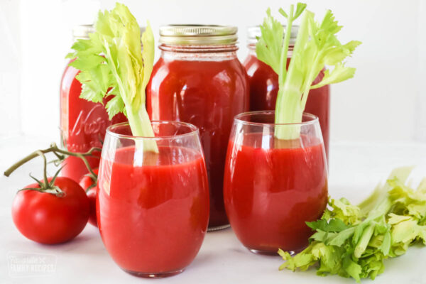 Two glasses of fresh tomato juice and canned tomato juice jars in the background.