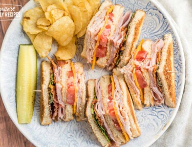 A club sandwich on a patterned plate with pickles and potato chips on the side
