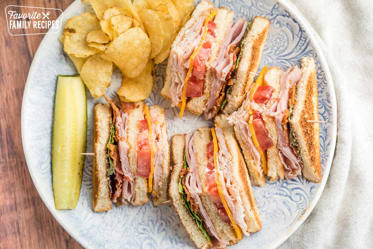 A club sandwich on a patterned plate with pickles and potato chips on the side