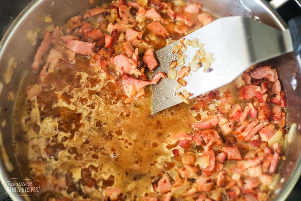 Bacon and onion in a skillet. The pan is being deglazed with a vinegar mixture and being stirred with a metal spatula.