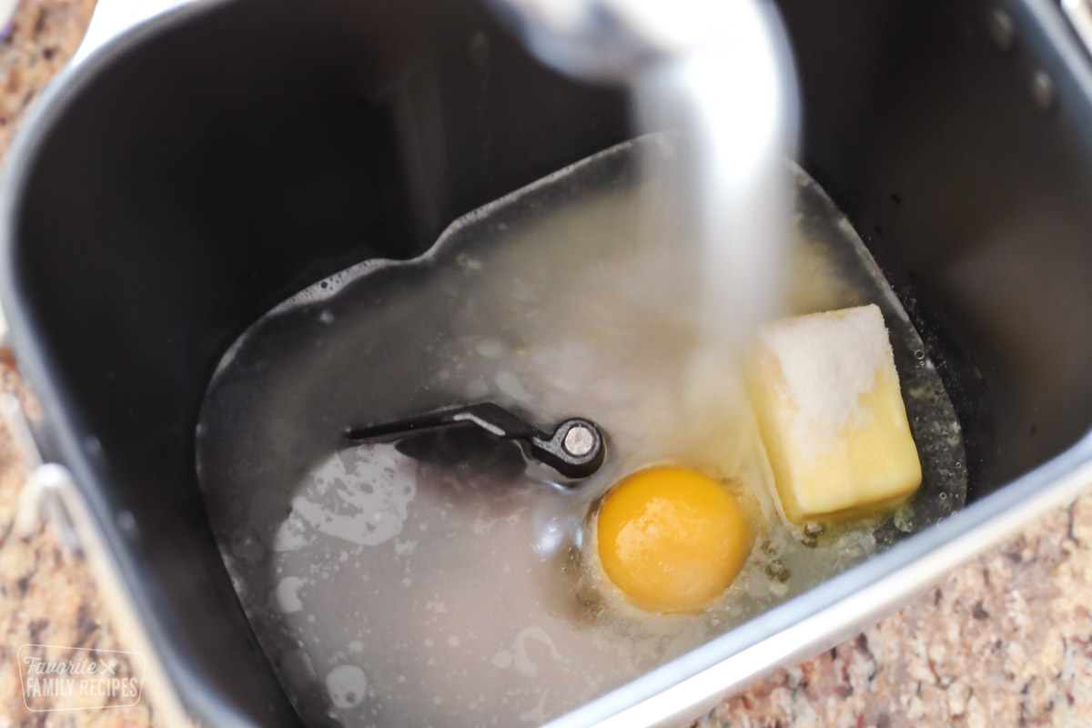 Sugar being poured into a bread machine along with an egg, butter, and water to make hamburger buns
