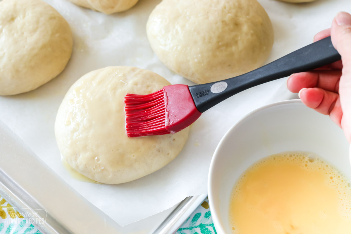An egg wash being brushed onto hamburger bun dough with a pastry brush.