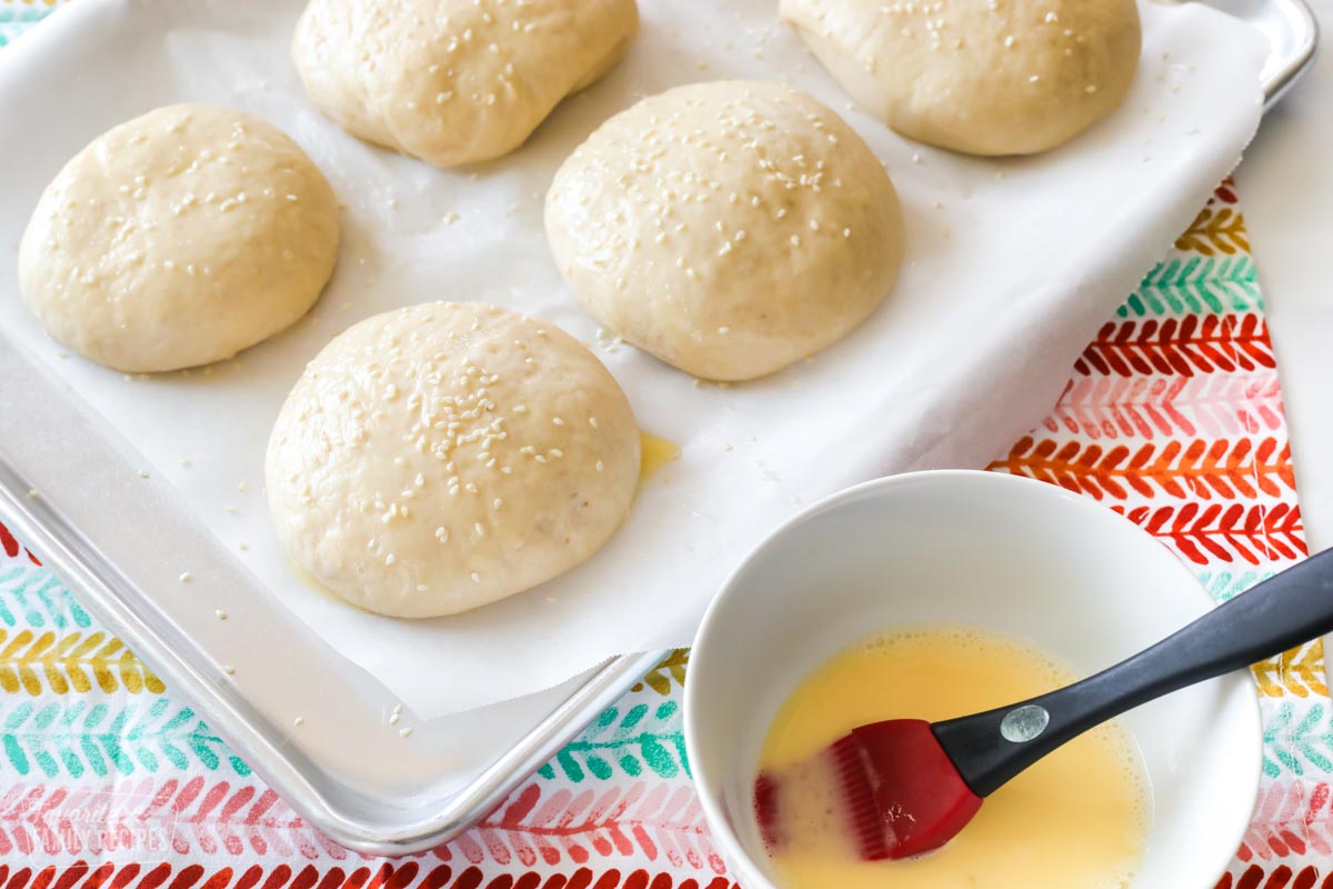 A baking sheet with dough formed into buns with an egg wash over the dough and sesame seeds sprinkled on top. A bowl of egg wash and a pastry brush are next to the baking sheet.