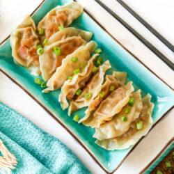 A rectangular plate full of potstickers sitting next to a dipping cup of potsticker sauce, chopsticks, and green onion.