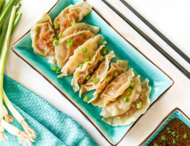 A rectangular plate full of potstickers sitting next to a dipping cup of potsticker sauce, chopsticks, and green onion.