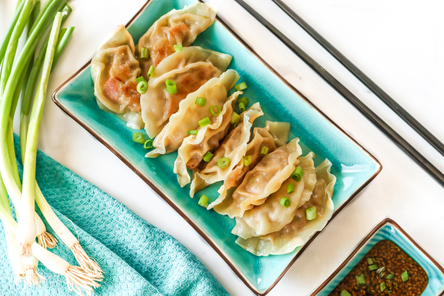 A rectangular plate full of potstickers sitting next to a dipping cup of potsticker sauce, chopsticks, and green onion.