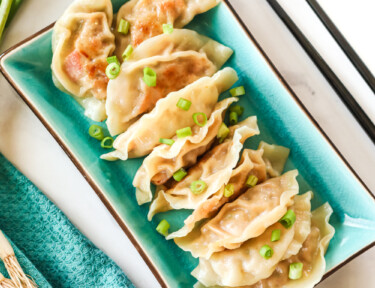 A rectangular plate containing potstickers with a garnish of green onion. It is sitting next to a cup of sauce and chopsticks.