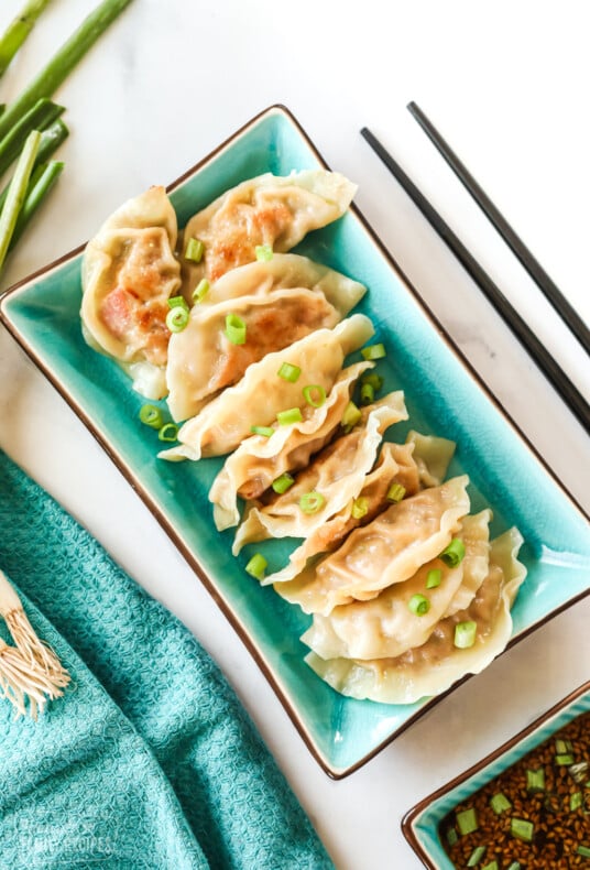 A rectangular plate containing potstickers with a garnish of green onion. It is sitting next to a cup of sauce and chopsticks.