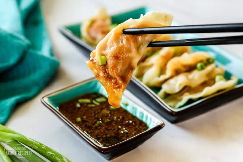 A potsticker being held with chopsticks dipped in soyaki sauce. A place of potstickers is in the background.