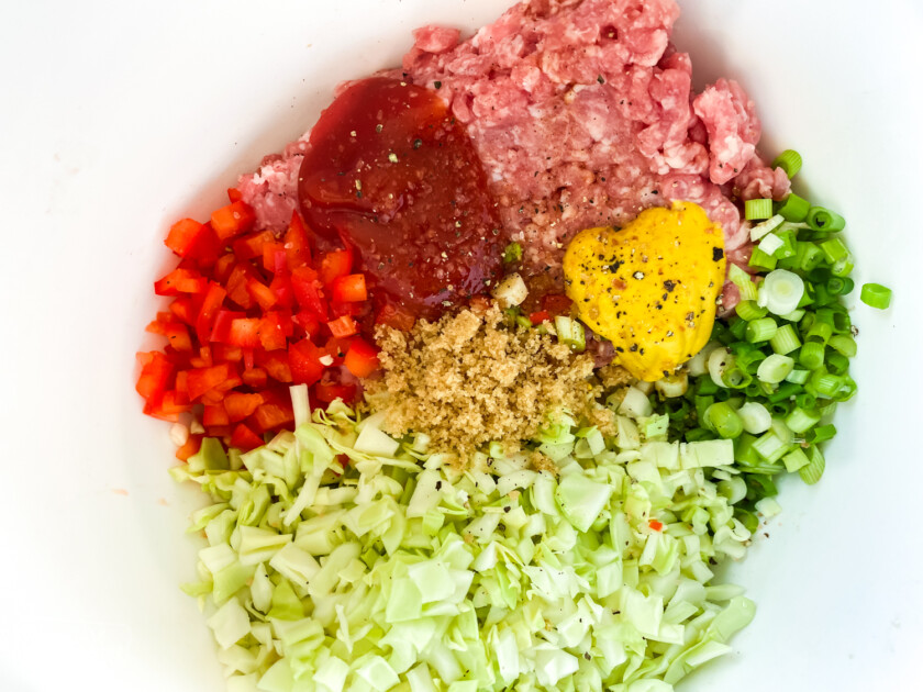 Ground pork, finely chopped cabbage, red bell peppers, green onion, ketchup, brown sugar, a beaten egg, salt, and pepper in a bowl ready to be mixed into a potsticker mixture.