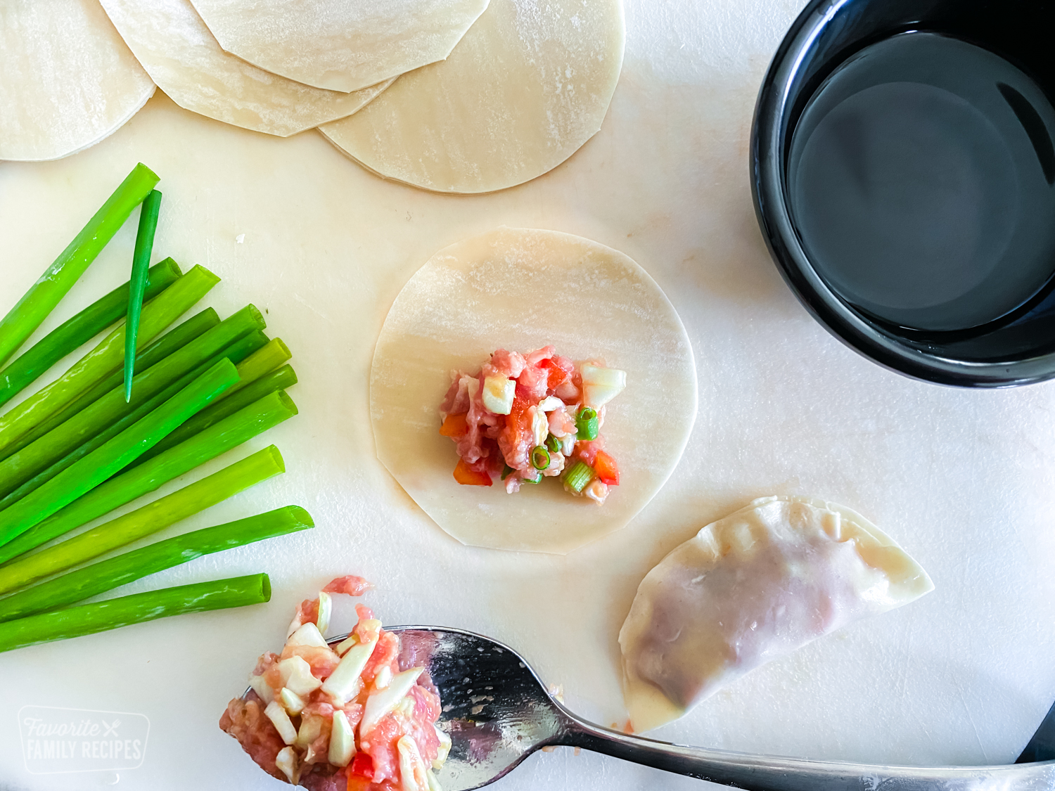 A round wonton wrapper with pork potsticker mixture placed in the middle. A completed potsticker, a spoon of mixture, a cup of water, and green onions are next to it.