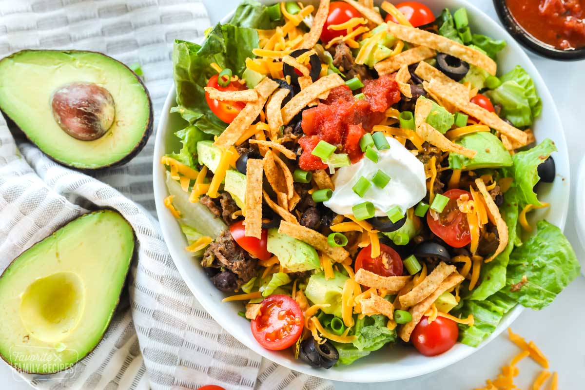 A taco salad made with ground beef, avocado, cheese, tomatoes, chips, salsa, and sour cream. The bowl is next to a halved avocado and a bowl of salsa.