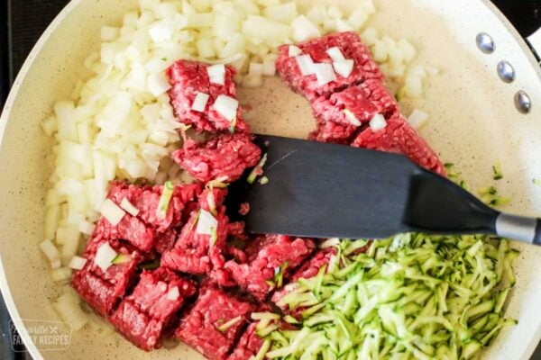 Uncooked ground beef, onion, and zucchini in a skillet with a spatula.