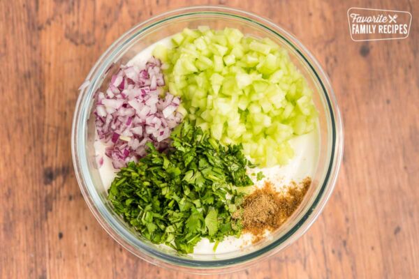 A glass bowl with yogurt, red onion, cucumber, cilantro, and cumin inside