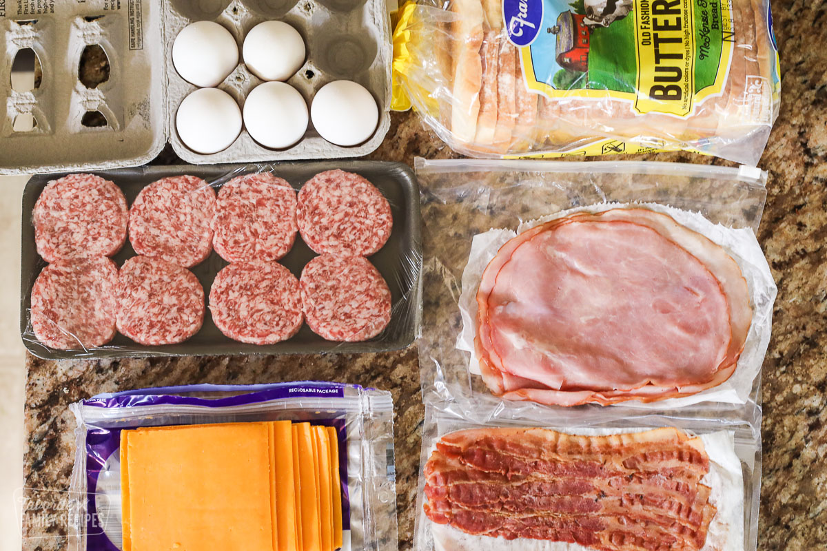 Ingredients to make a breakfast sandwich laid out on a counter including sausage, cheese, bread, bacon, and ham