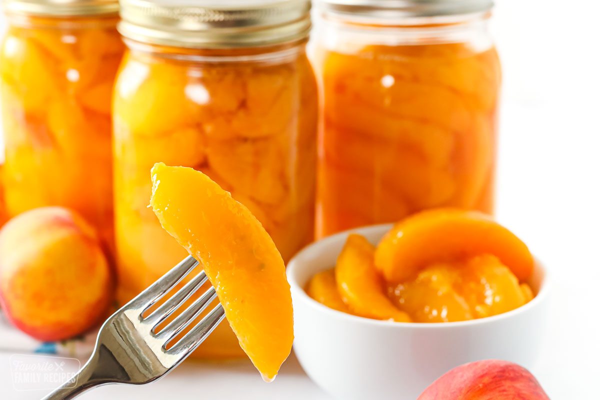 A close up of a peach slice on a fork in front of bottled peaches