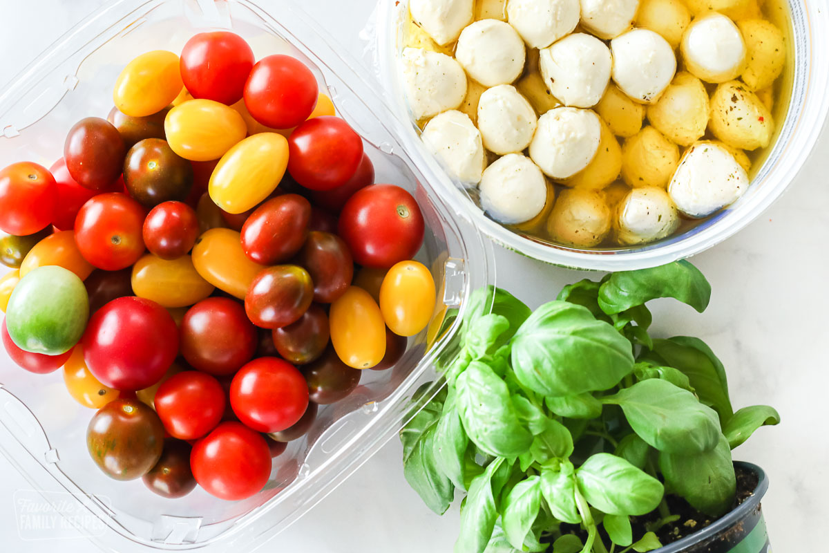 A carton of cherry tomatoes, a container of mozzarella balls, and a basil plant.