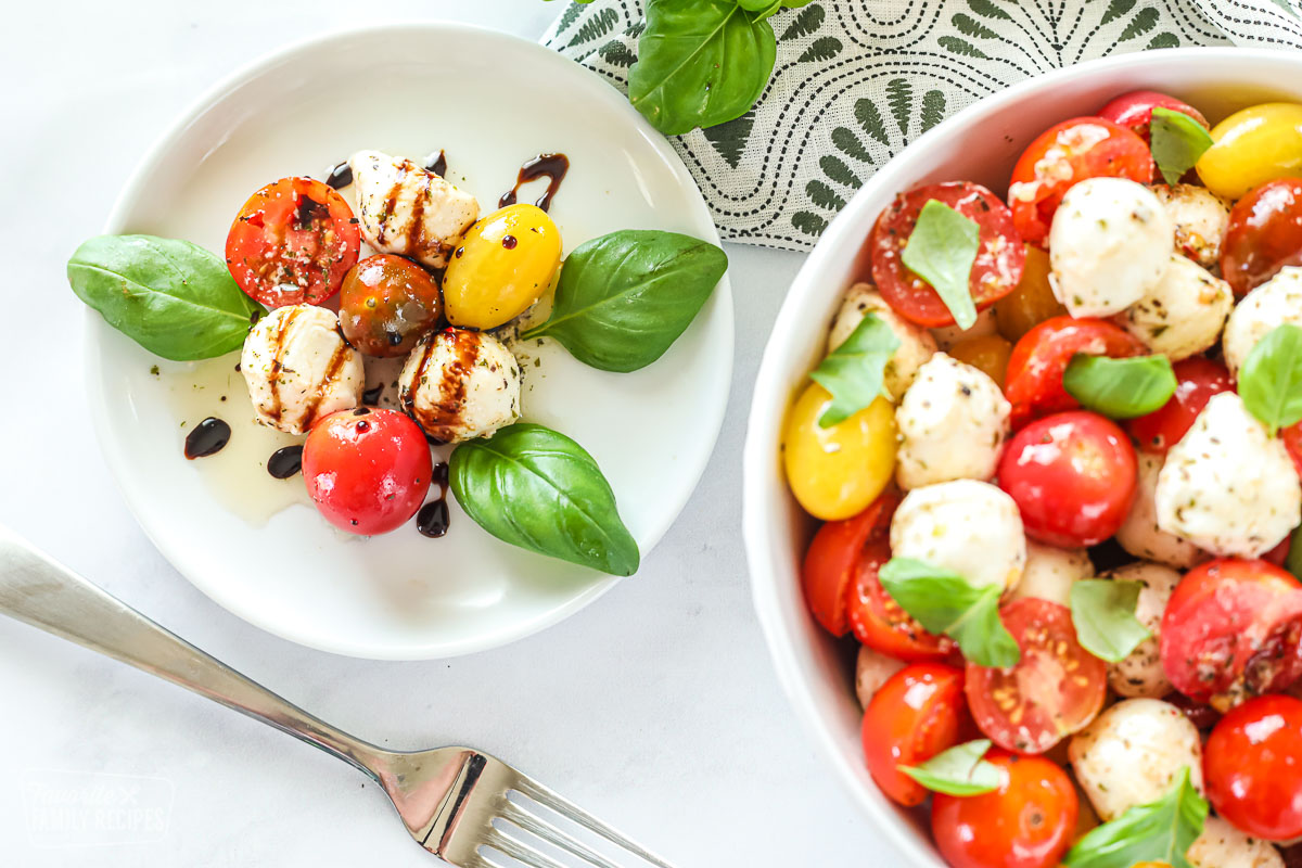 A small plate of cherry tomatoes and baby mozzarella next to a large bowl of the same. Both are garnished with basil.