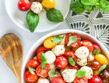 A small plate of cherry tomatoes, baby mozzarella, and basil leaves next to a large bowl of the same.