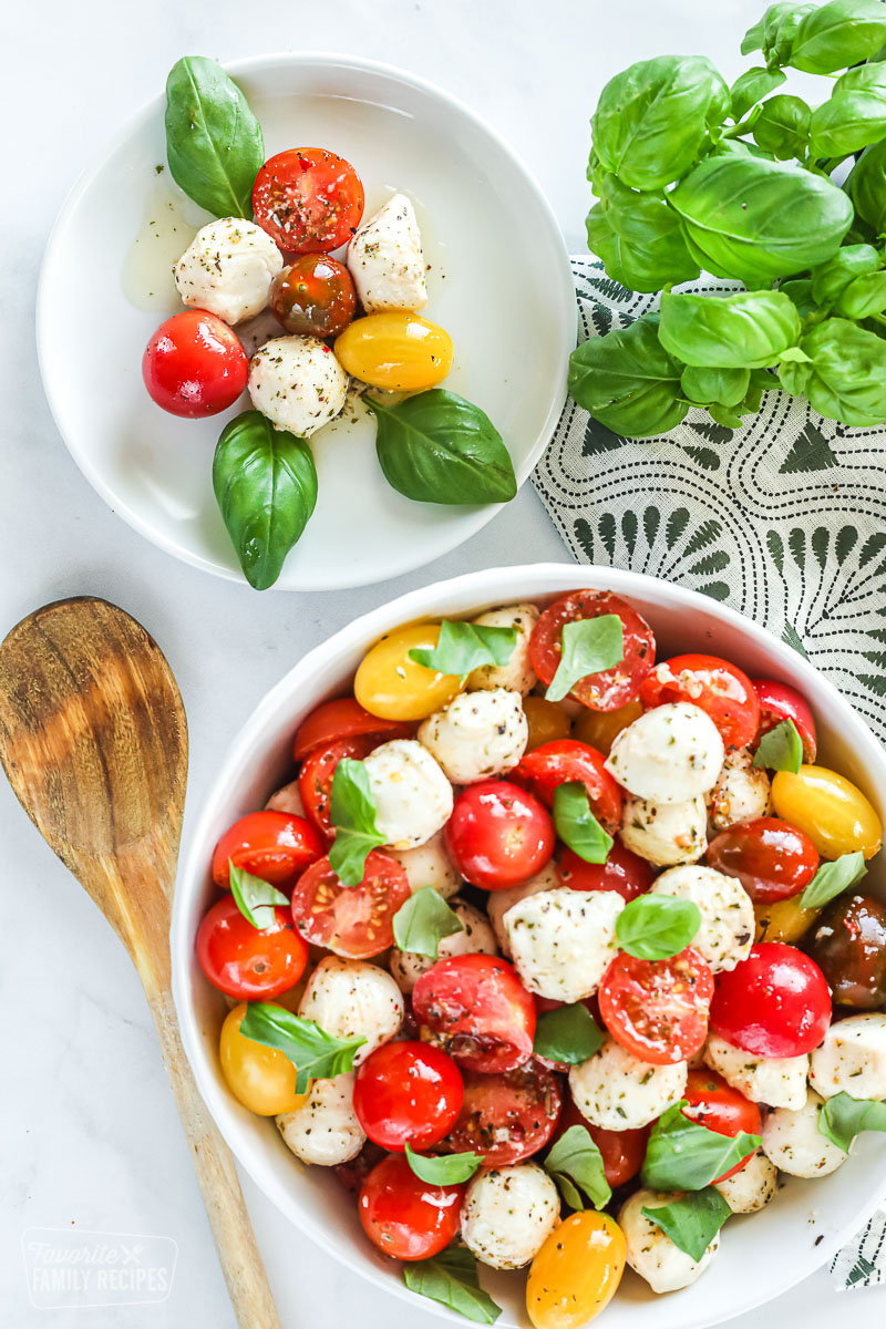 A small plate of cherry tomatoes, baby mozzarella, and basil leaves next to a large bowl of the same.