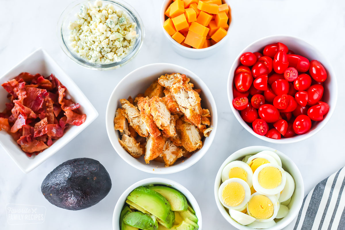 Bowls of salad toppings including tomatoes, egg, avocado, bacon, blue cheese, and chicken.