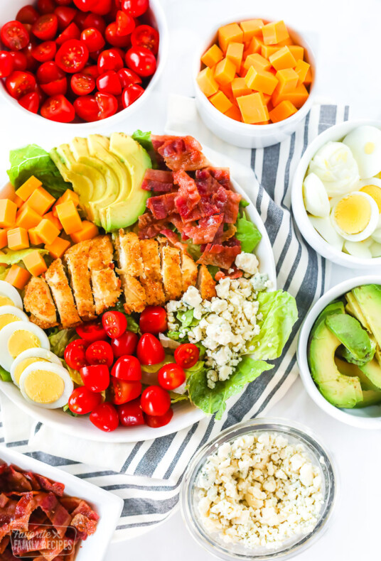 A bowl of Cobb salad with toppings surrounding the bowl including bacon, blue cheese, avocado, egg, cheese, and tomatoes.