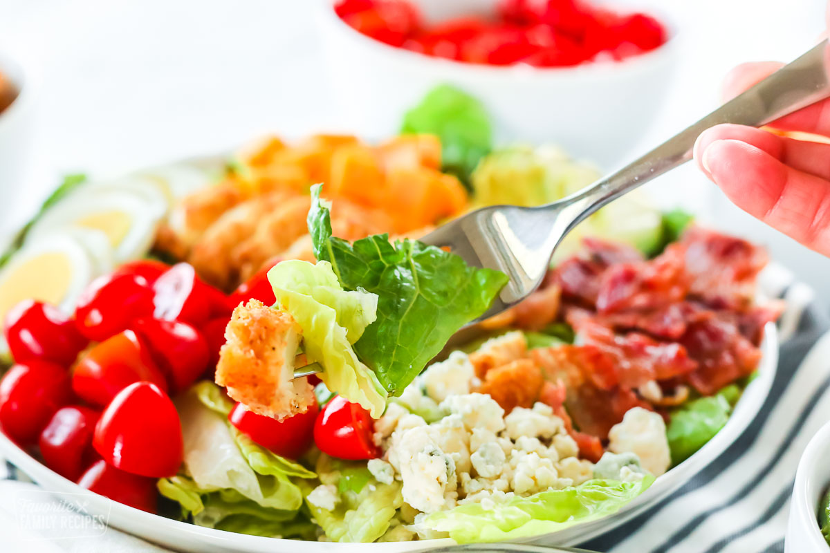 A fork holding lettuce and a piece of cooked chicken over a bowl of Cobb salad