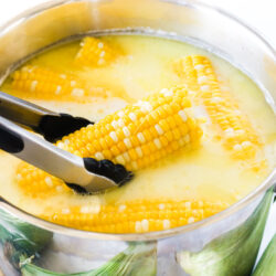 Corn on the cob in a stock pot. One piece is being puled out with a pair of tongs.