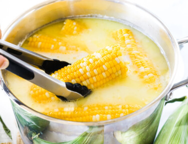 Corn on the cob in a stock pot. One piece is being puled out with a pair of tongs.