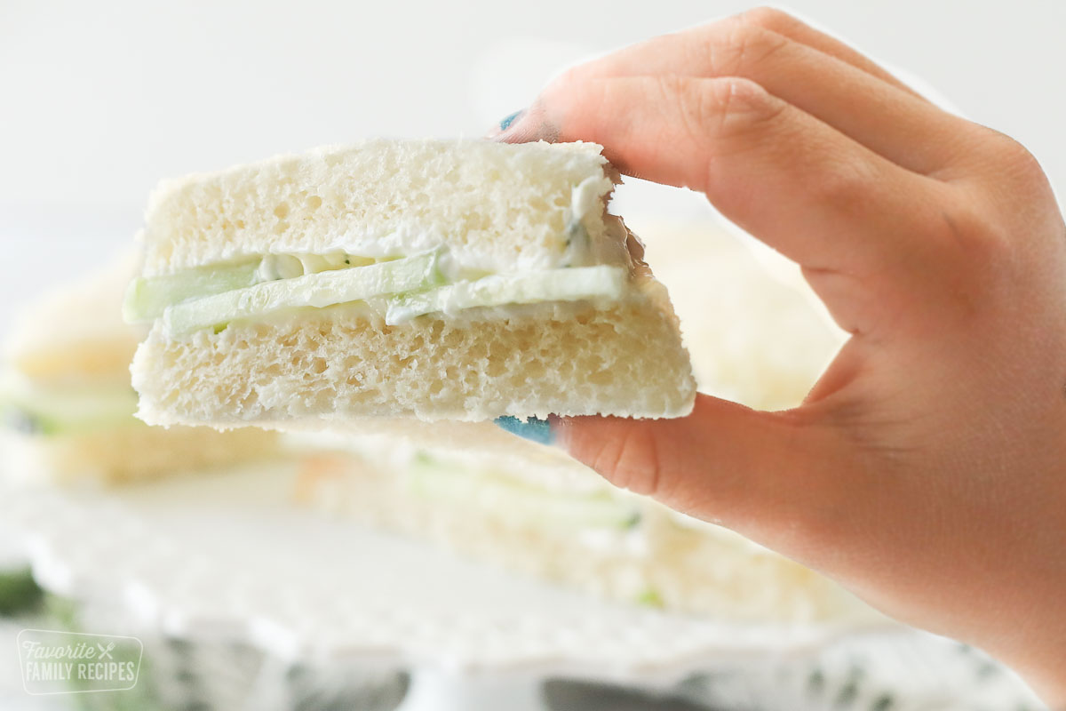 A cucumber tea sandwich wedge being help above a tray of cucumber sandwiches.