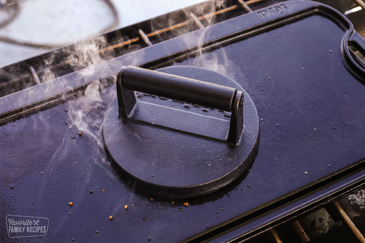 A cast iron burger press pressing down on a hamburger over the grill