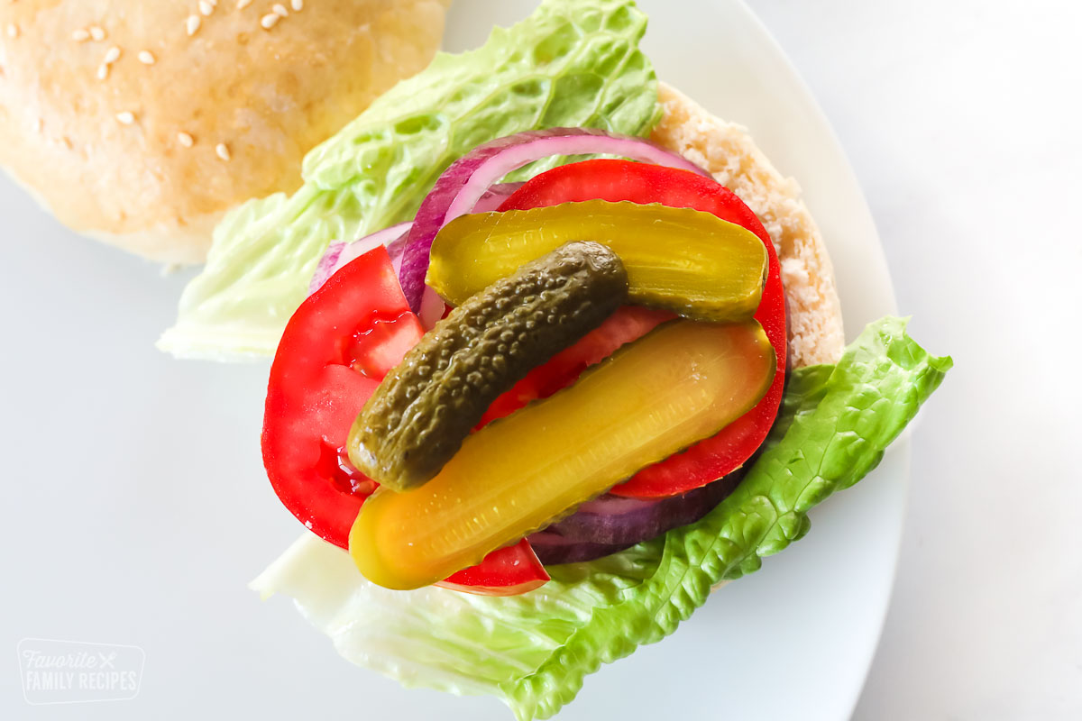 An open faced hamburger bun layered with lettuce, tomatoes, pickles and sliced onion over the top