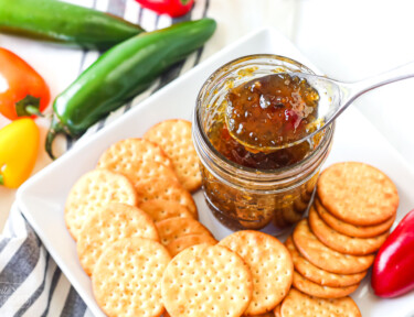 A spoon taking a scoop out of a jar of jalapeño jelly on a plate with crackers