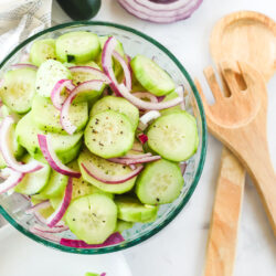 Cucumbers in a bowl marinated with red onion. Wooden spoons, a sliced onion, and a plate are next to the bowl
