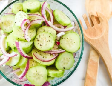 Cucumbers in a bowl marinated with red onion. Wooden spoons, a sliced onion, and a plate are next to the bowl