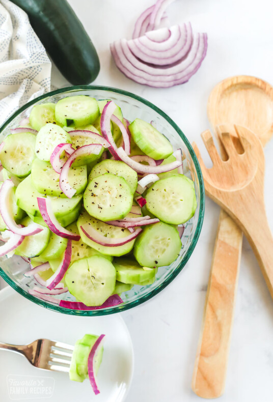 Cucumbers in a bowl marinated with red onion. Wooden spoons, a sliced onion, and a plate are next to the bowl