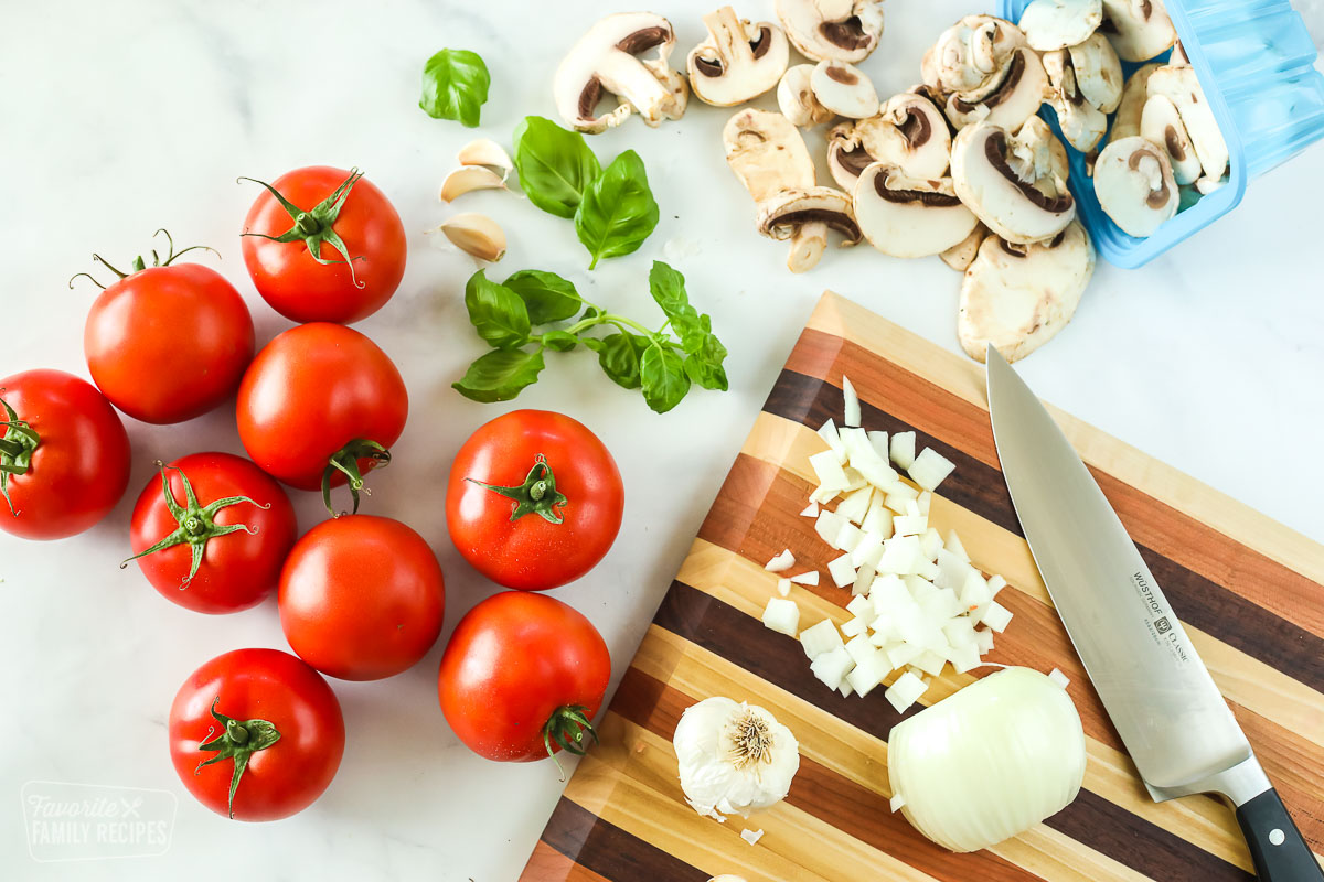 Fresh tomatoes, basil, garlic and mushrooms next to a cutting board with an onion and a knife