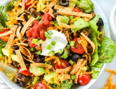 A taco salad made with ground beef, avocado, cheese, tomatoes, chips, salsa, and sour cream. The bowl is next to a halved avocado, cherry tomatoes, and a bowl of salsa.