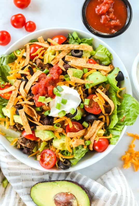 A taco salad made with ground beef, avocado, cheese, tomatoes, chips, salsa, and sour cream. The bowl is next to a halved avocado, cherry tomatoes, and a bowl of salsa.