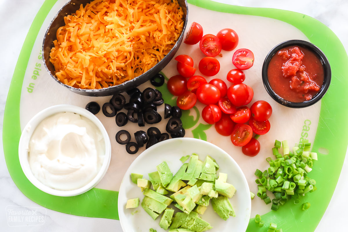 A container of shredded cheese, a cup of sour cream, a cup of salsa, diced avocado, sliced olives, green onion, and cherry tomatoes on a cutting board.