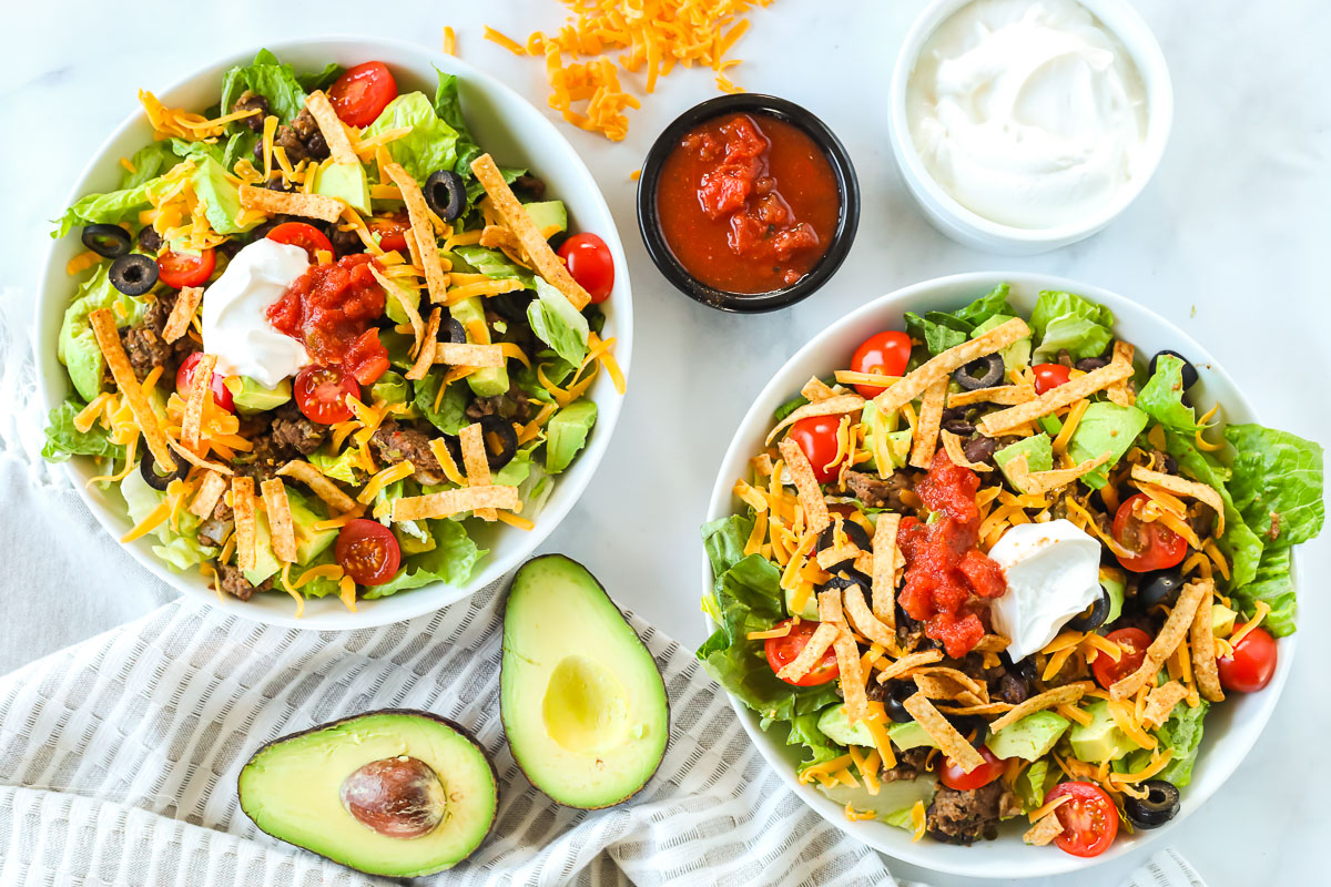 Two bowls of taco salad. An avocado, a cup of sour cream, and a cup of salsa are sitting next to the bowls.
