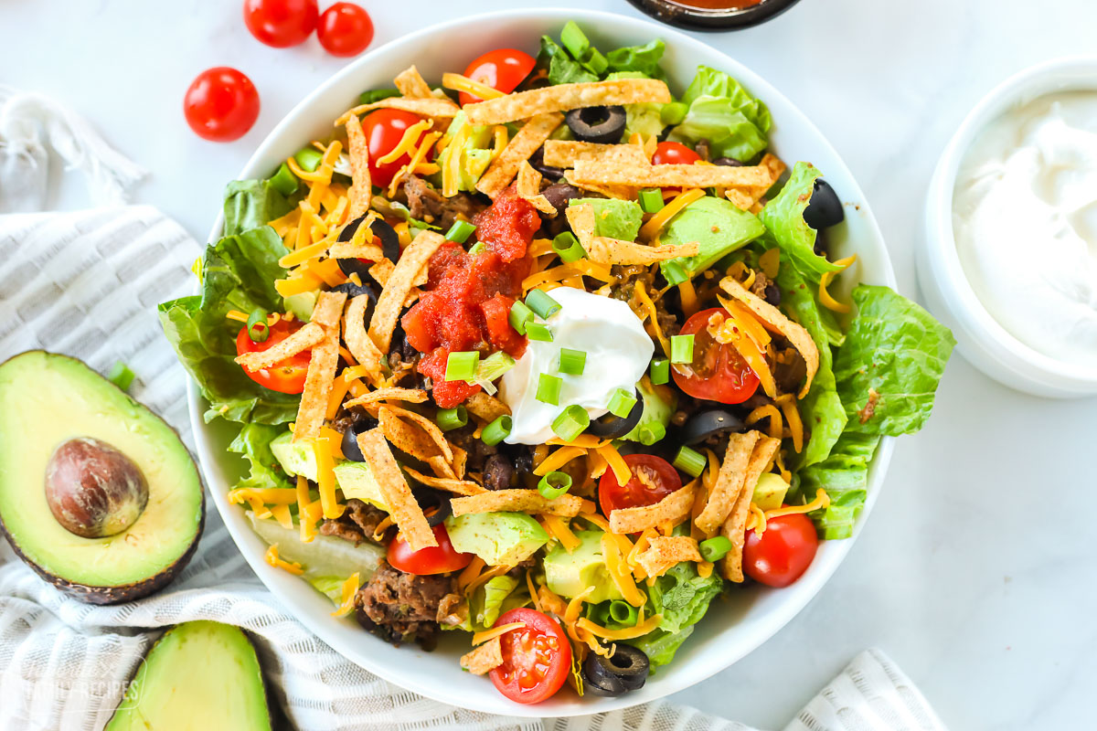 A taco salad made with ground beef, avocado, cheese, tomatoes, chips, salsa, and sour cream. The bowl is next to a halved avocado, cherry tomatoes, a cup of sour cream, and a bowl of salsa.